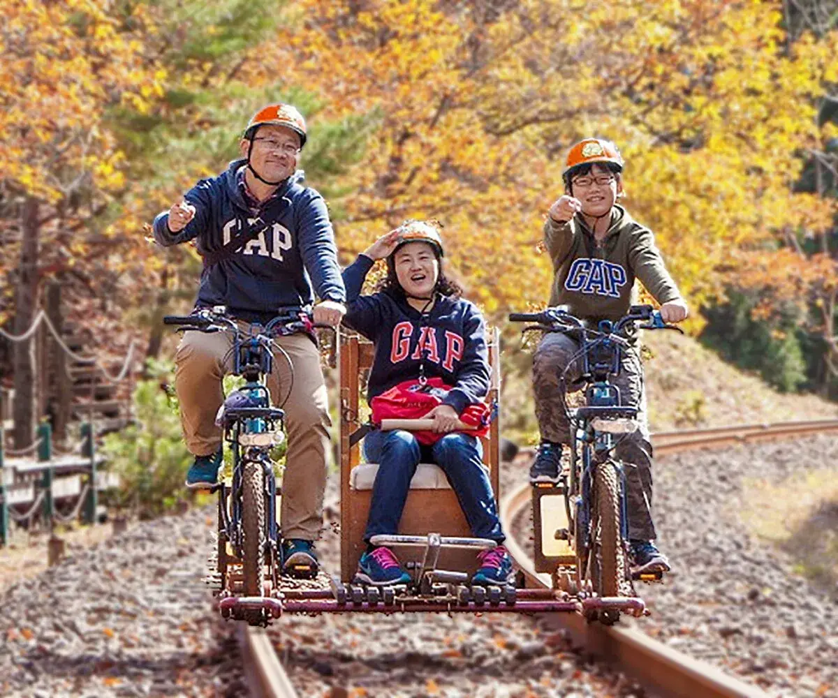 Family on a central-bench rail bike: two pedalers and one passenger on the wooden seat, autumn in Kamioka
