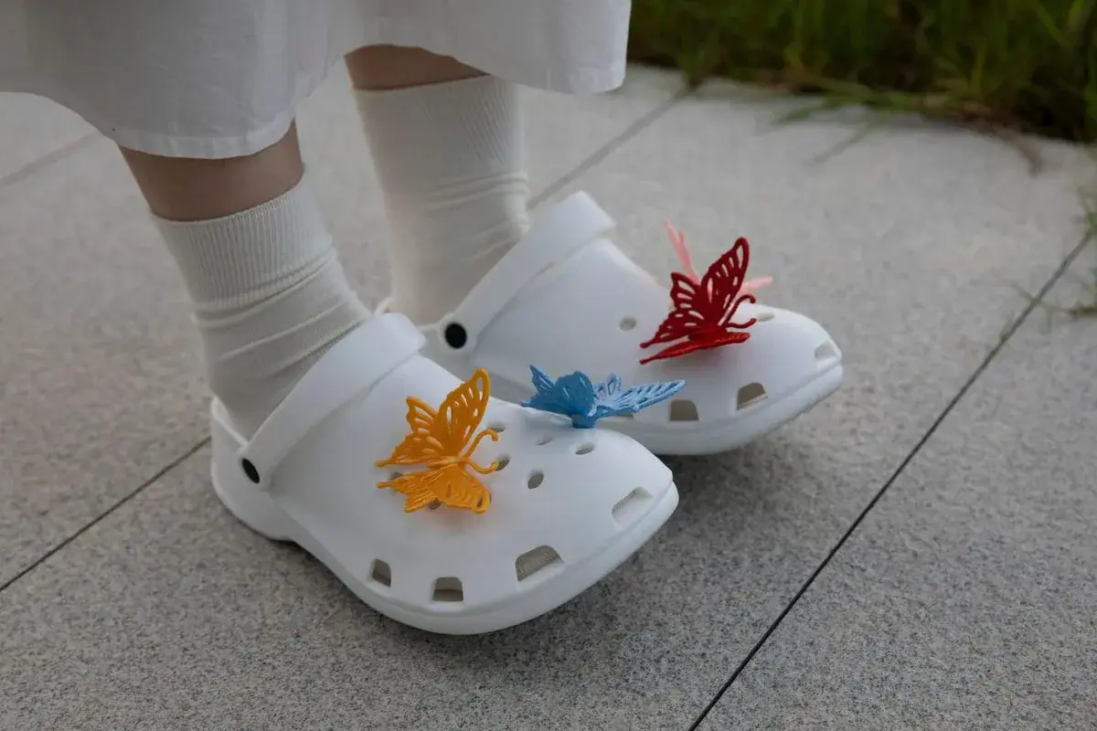 Person walking outdoors wearing white 3D-printed clogs decorated with colorful butterfly charms